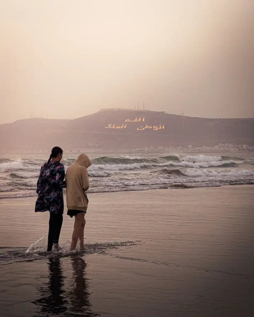 Two people walking on Agadir beach at sunset with the Oufella hill inscription behind